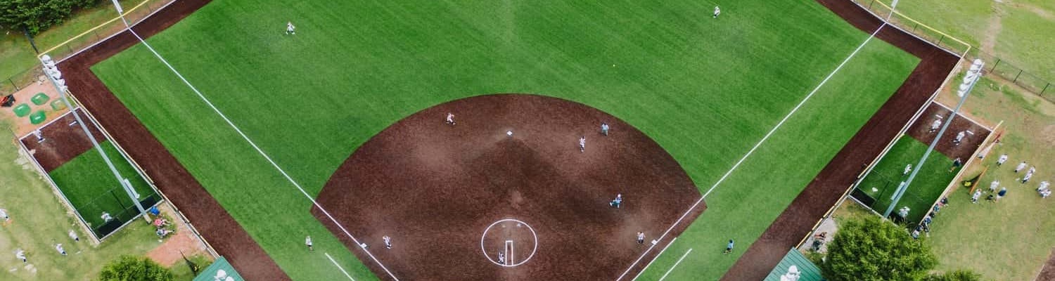 A young baseball player in a black shirt and gray pants pitches from a red mound on a green artificial turf installation sports field, with teammates and buildings in the background, possibly in Salt Lake City.