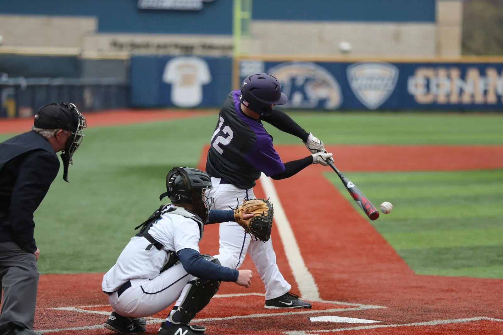 A baseball player in a purple and black uniform swings at a pitch on an outdoor field with sports field artificial turf, while a catcher and umpire watch closely behind home plate.