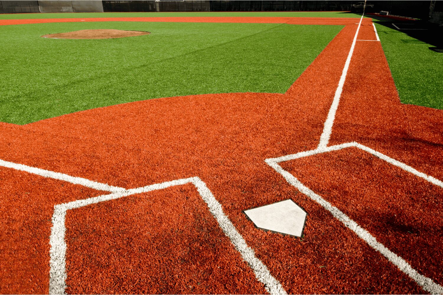 A young baseball player in a black shirt and gray pants pitches from a red mound on a green artificial turf installation sports field, with teammates and buildings in the background, possibly in Salt Lake City.