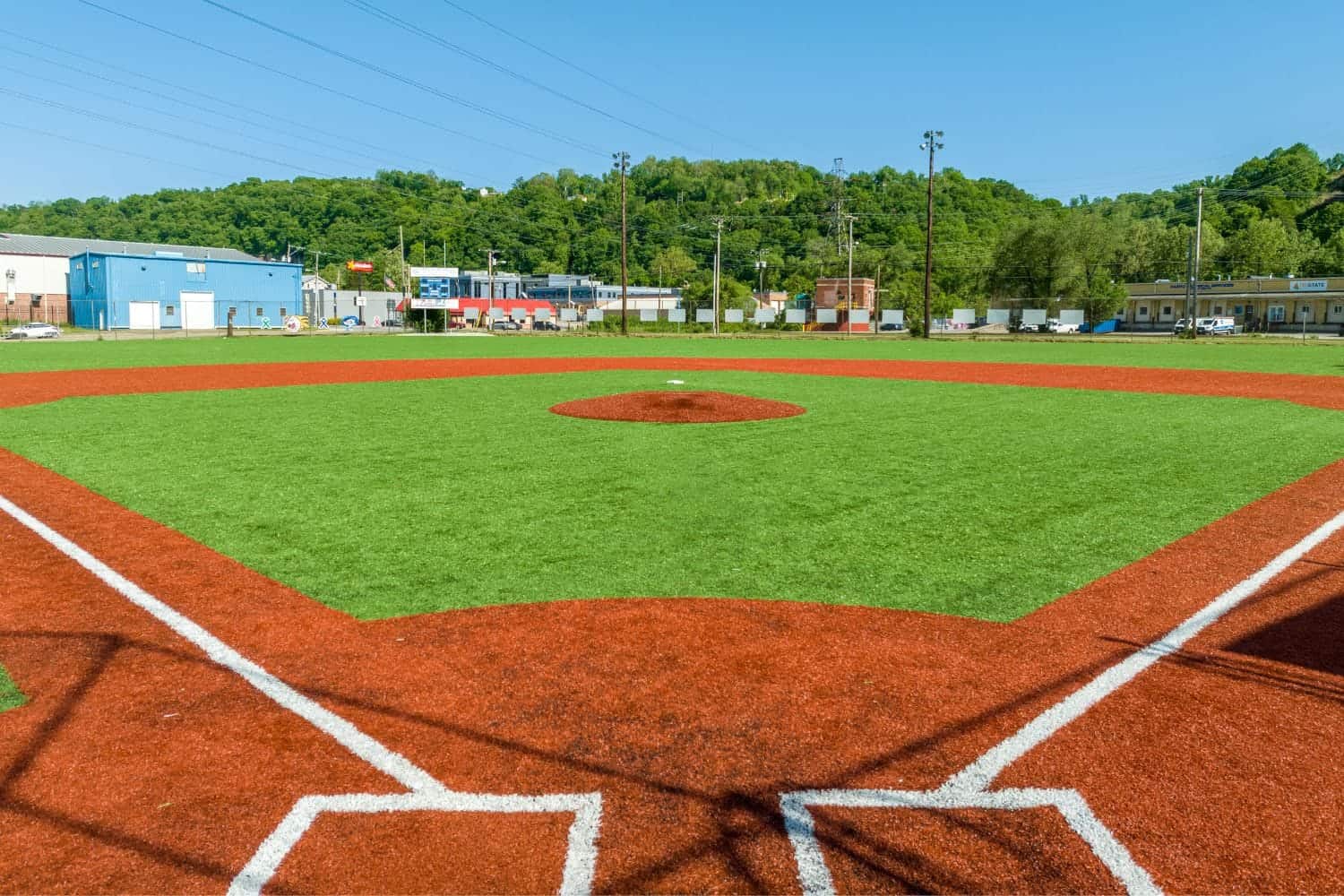 A young baseball player in a black shirt and gray pants pitches from a red mound on a green artificial turf installation sports field, with teammates and buildings in the background, possibly in Salt Lake City.