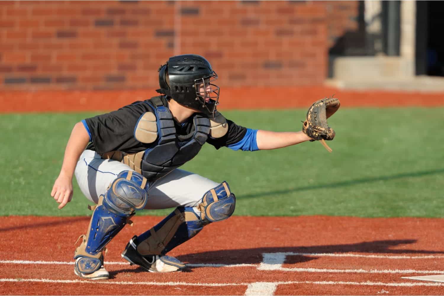 A young baseball player in a black shirt and gray pants pitches from a red mound on a green artificial turf installation sports field, with teammates and buildings in the background, possibly in Salt Lake City.