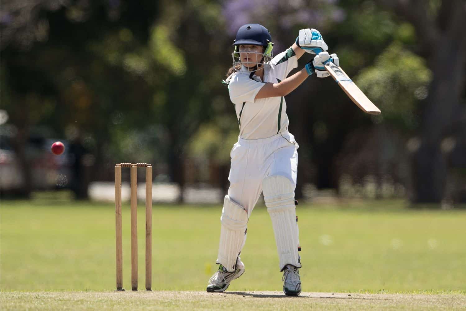 A young cricket player in protective gear prepares to hit a ball with a bat on sports turf. The ball is mid-air, and a set of stumps stands behind the player. Trees are blurred in the background.