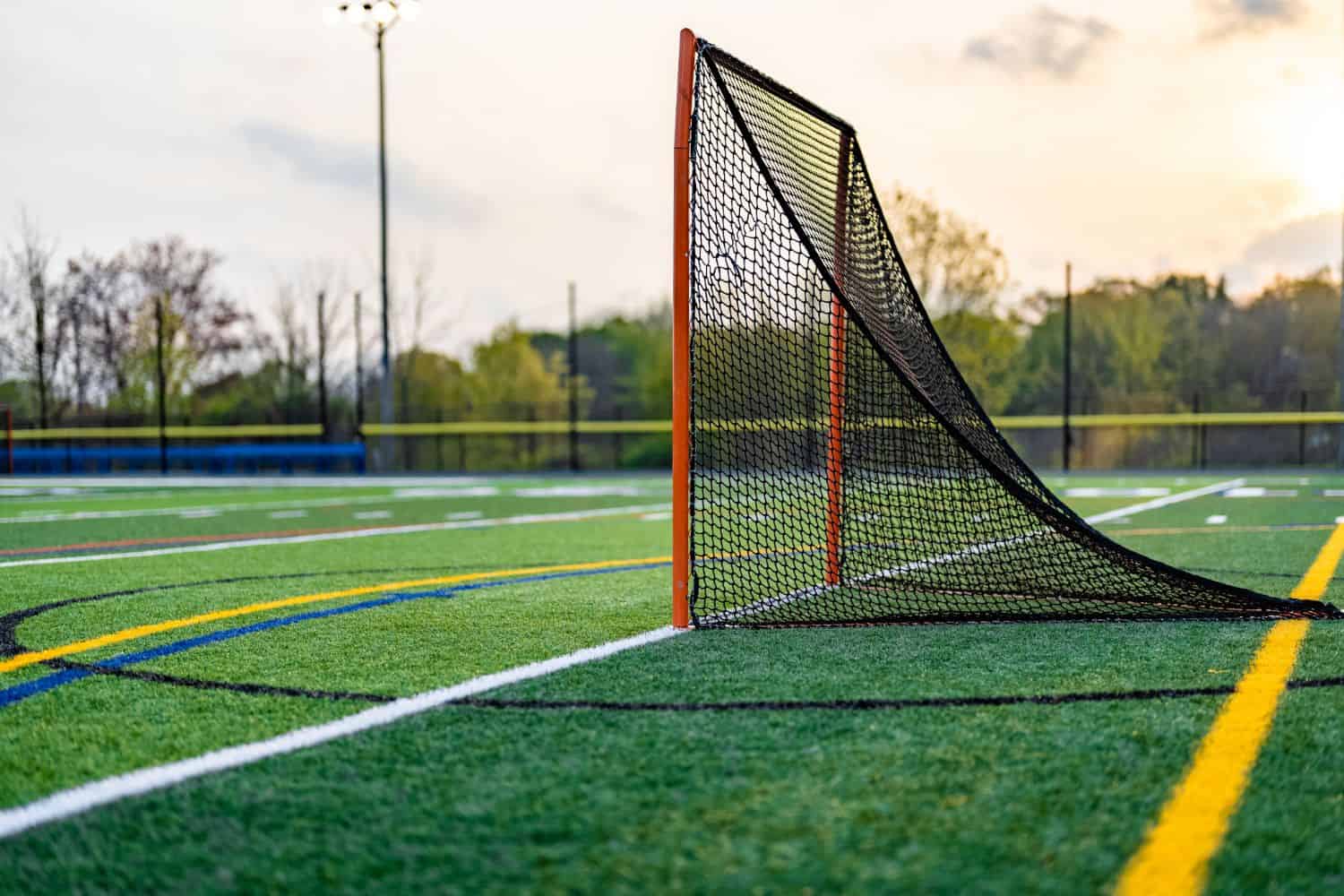 A lacrosse goal net stands on a green artificial turf field, showcasing expert artificial turf installation, with colorful lines for various sports fields, a running track, and bleachers visible in the background.