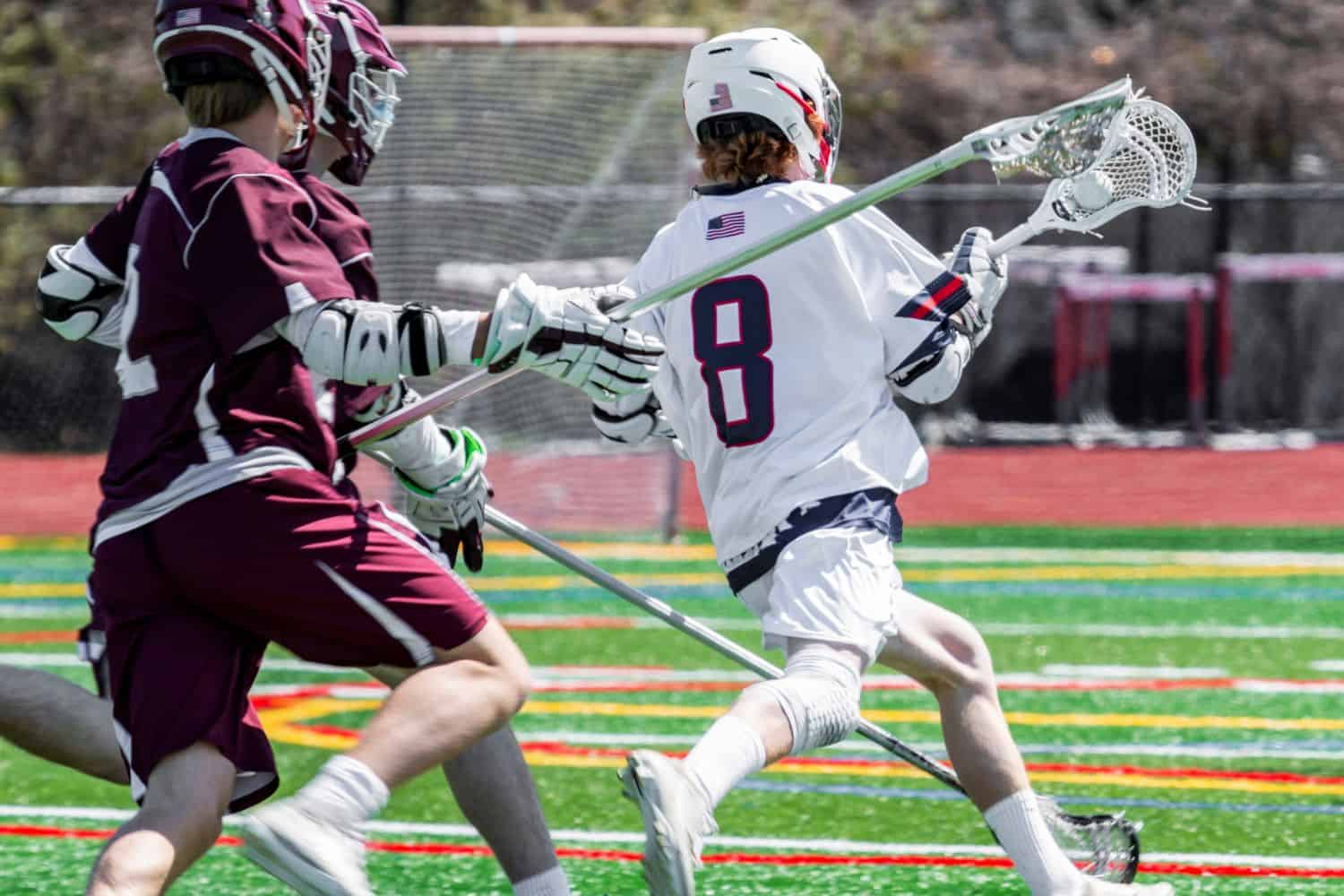 Two lacrosse players in protective gear compete on an artificial turf sports field. Number 8 in white runs with the ball as a maroon-clad player defends closely, all under the bright Phoenix sun.