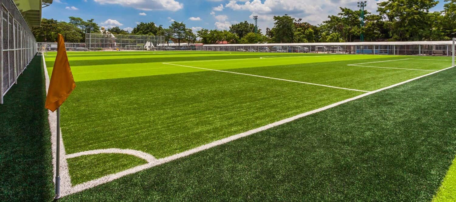 A rugby ball rests on synthetic turf with crisp white markings, as goalposts and tall trees stand in the blurred background of Albuquerque’s sports fields on a sunny day.