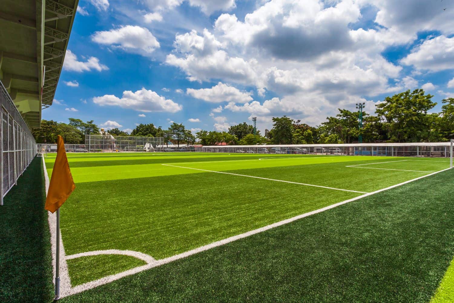 A bright, sunny day over an empty artificial turf sports field in Phoenix AZ, with clear white lines, a yellow corner flag, and trees and buildings in the background under a partly cloudy blue sky.