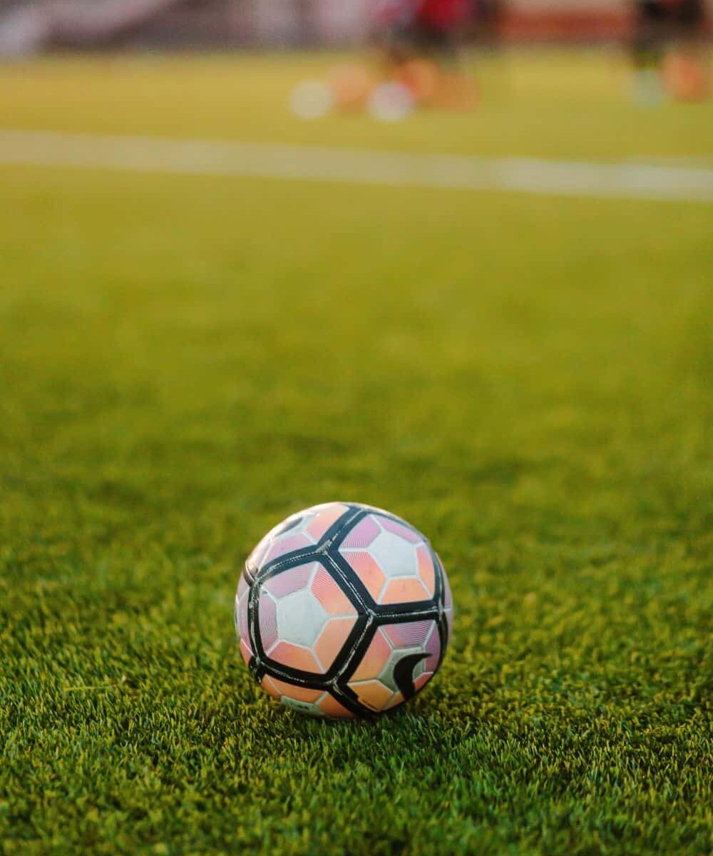 A close-up view of a corner of a soccer field in Irvine CA at dusk, with bright stadium lights illuminating the artificial turf and a fence in the background.