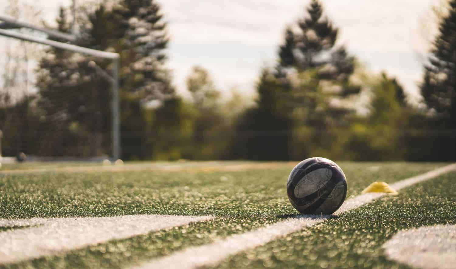 A rugby ball rests on synthetic turf with crisp white markings, as goalposts and tall trees stand in the blurred background of Albuquerque’s sports fields on a sunny day.