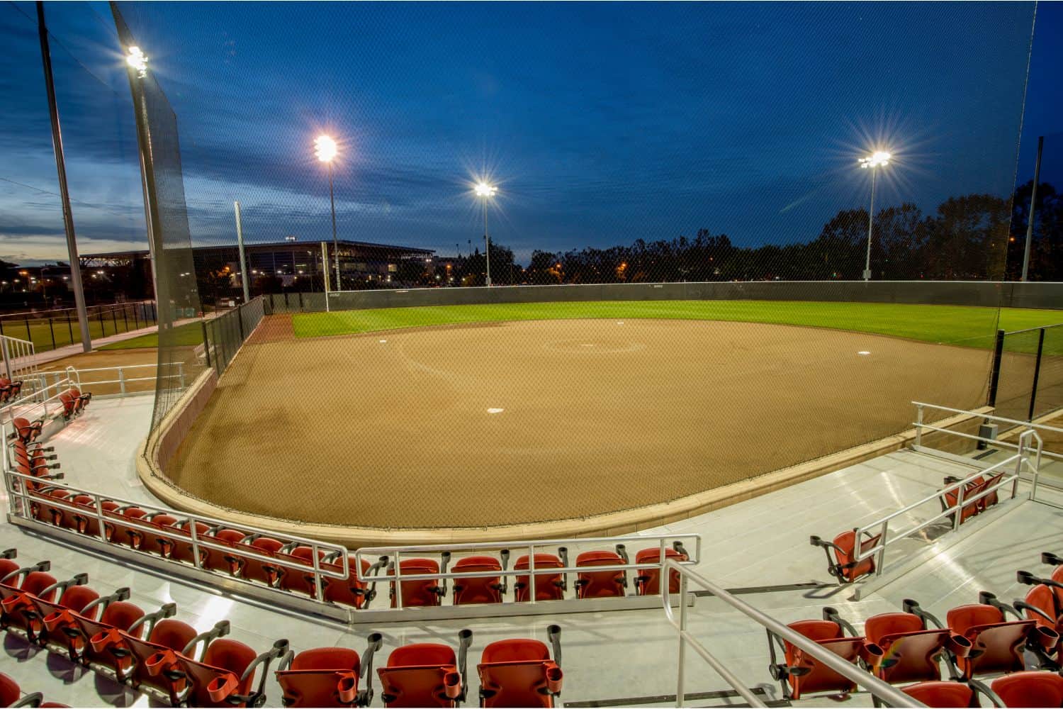 A well-lit, empty baseball or softball field with sports turf viewed from the stands at dusk, featuring red seats in the foreground and a green outfield surrounded by a safety net.