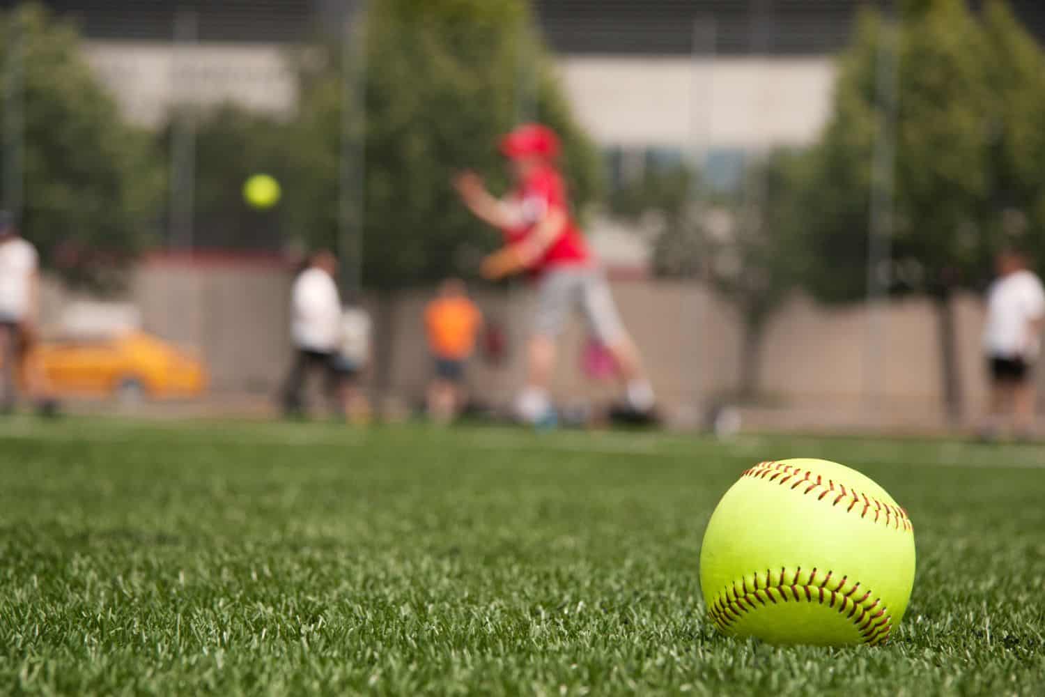 A yellow softball lies on green grass in the foreground of a sports field, with a blurred scene of people playing softball in the background.