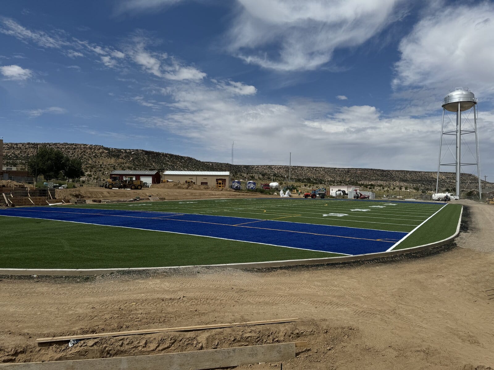 A young cricket player in protective gear prepares to hit a ball with a bat on sports turf. The ball is mid-air, and a set of stumps stands behind the player. Trees are blurred in the background.