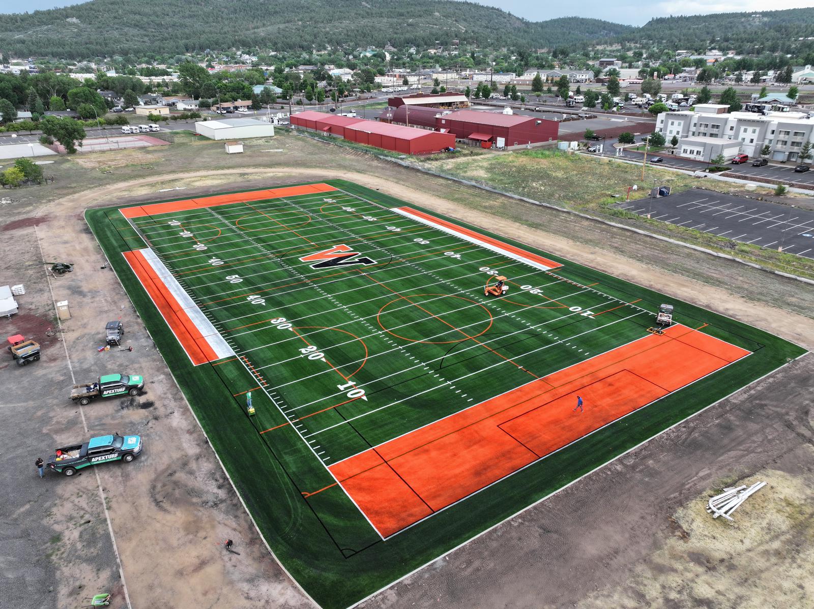 Aerial view of a football field with bright green artificial turf. “FALCONS” is written in both end zones, and “CACTUS SHADOWS” is along the sides. The sports field features white yard lines, numbers, and blue accents.
