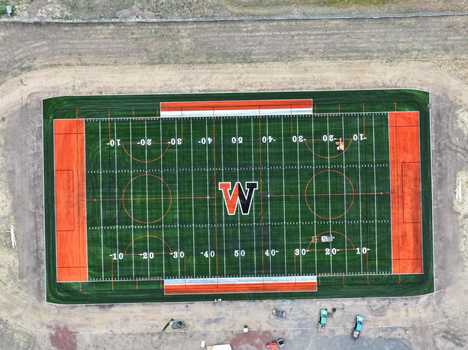 Aerial view of an artificial turf sports field in Irvine CA with a large W at center, orange end zones, green stripes, yard lines, and various field markings. Construction equipment is visible nearby.