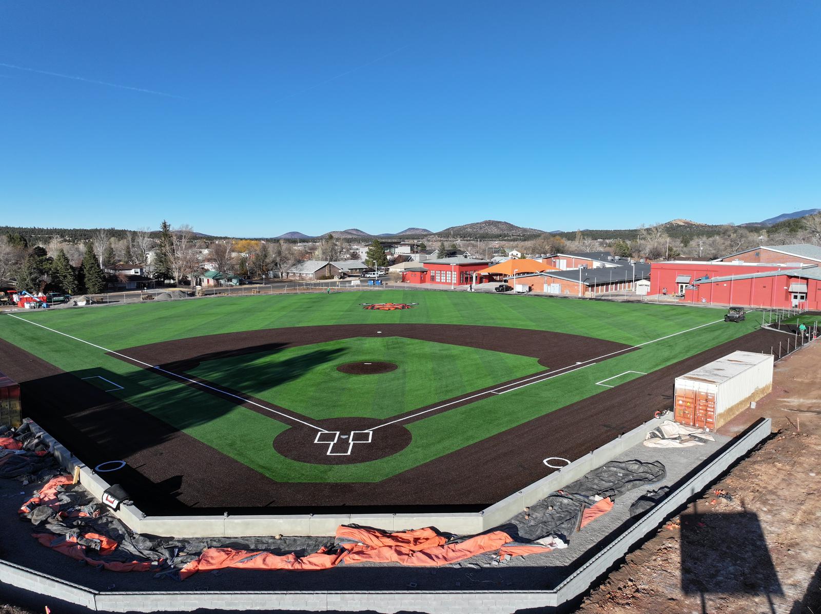 A young baseball player in a black shirt and gray pants pitches from a red mound on a green artificial turf installation sports field, with teammates and buildings in the background, possibly in Salt Lake City.